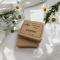 Calamine goat milk soap bars with kraft paper labels styled with fresh white daisies, dappled sunlight, and soft linen on a bright white surface