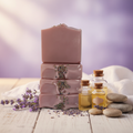 Stacked lavender goat milk soap bars styled with fresh lavender sprigs, linen, and essential oil bottles on a rustic white wood surface