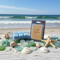 Coastal Waters wax melt clamshell packages displayed on weathered driftwood with sea glass, seashells, a starfish, and soft sand with ocean waves in the background