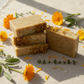 Calendula goat milk soap bars topped with dried golden calendula petals, styled on natural linen with fresh calendula flowers and herb sprigs