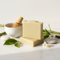 Green tea goat milk soap bars styled on white marble with fresh green tea leaves, ceramic matcha bowl, bamboo, and smooth stones